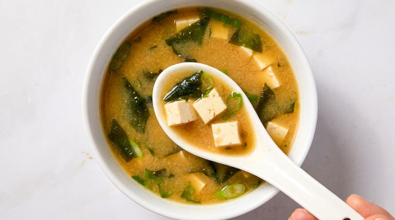 Overhead view of miso soup with tofu, seaweed, and scallions in a white bowl, with a spoon lifting some.