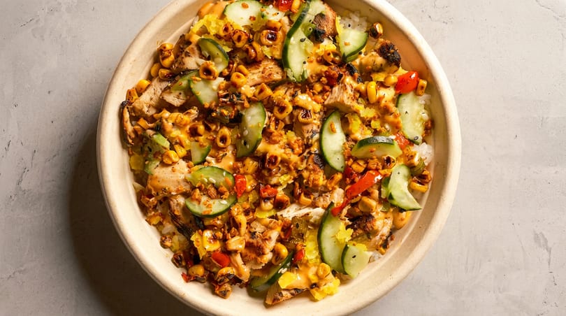 Overhead view of a bowl with grilled chicken, corn, cucumbers, and red peppers over rice.