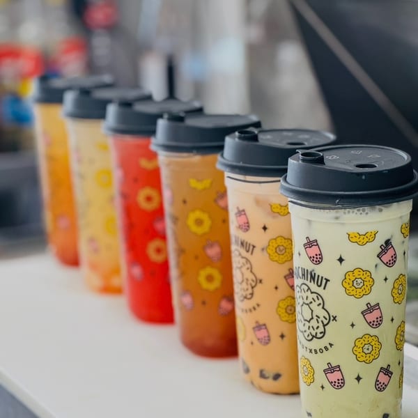 Six vibrant Mochinut boba tea drinks with fruit and donut patterns, lined on a counter.
