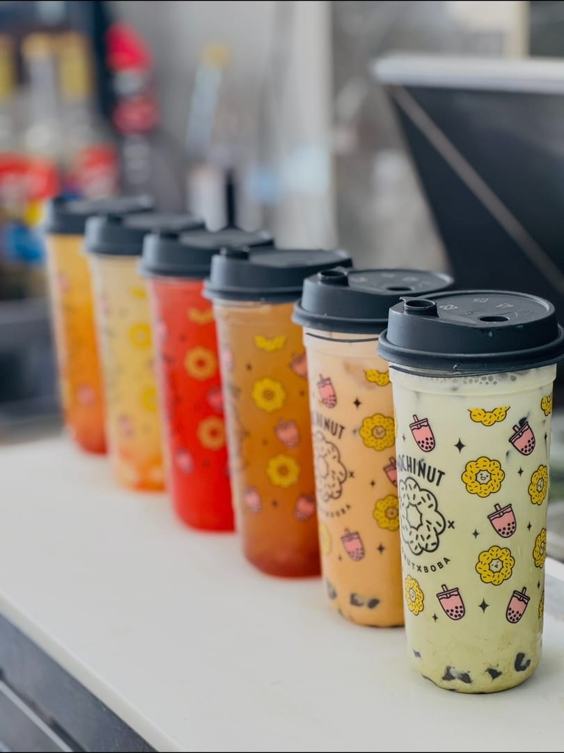 Six vibrant Mochinut boba tea drinks with fruit and donut patterns, lined on a counter.