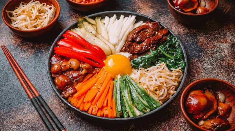 Colorful Korean bibimbap bowl with beef, vegetables, and egg yolk, with chopsticks and side dishes.