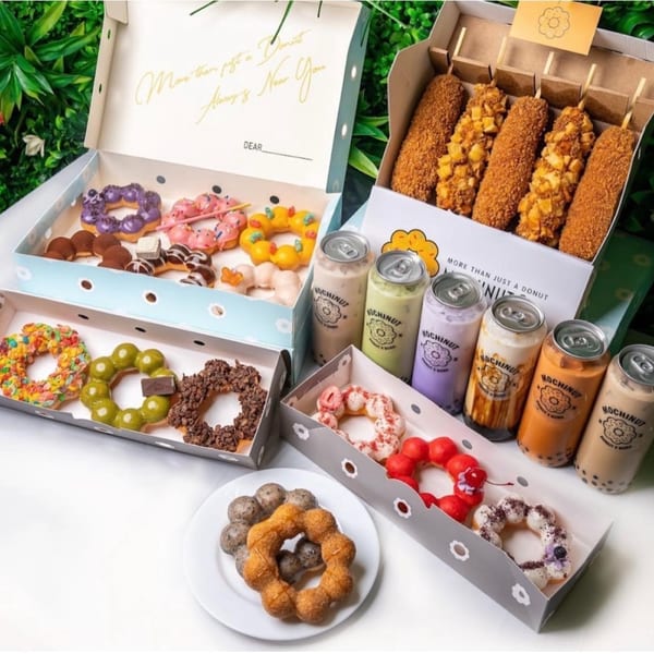 Assortment of colorful Mochinut donuts, corn dogs, and canned bubble tea drinks on a white surface.
