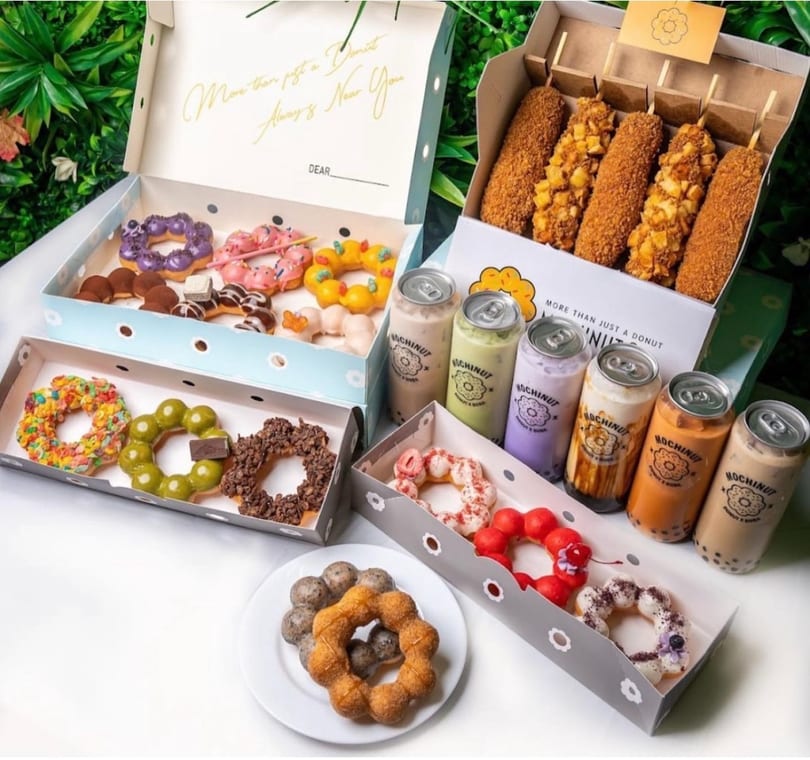 Assortment of colorful Mochinut donuts, corn dogs, and canned bubble tea drinks on a white surface.