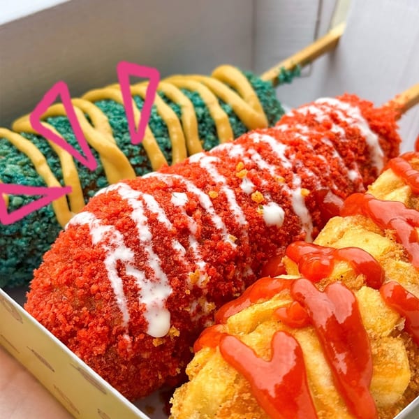Two colorful corn dogs and fried potatoes with sauces in a white takeaway box.