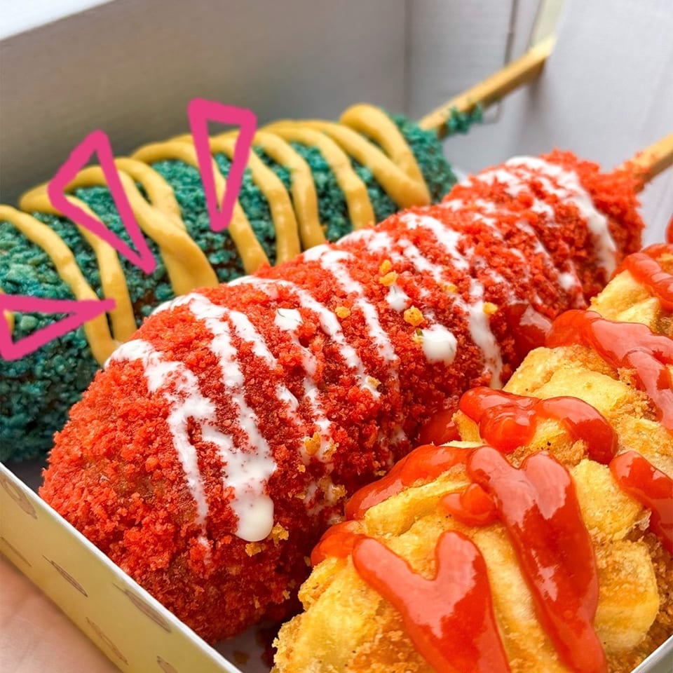 Two colorful corn dogs and fried potatoes with sauces in a white takeaway box.