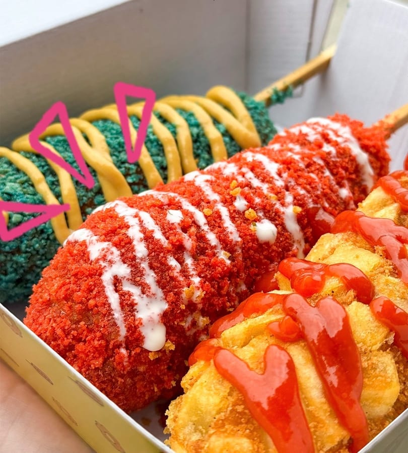 Two colorful corn dogs and fried potatoes with sauces in a white takeaway box.