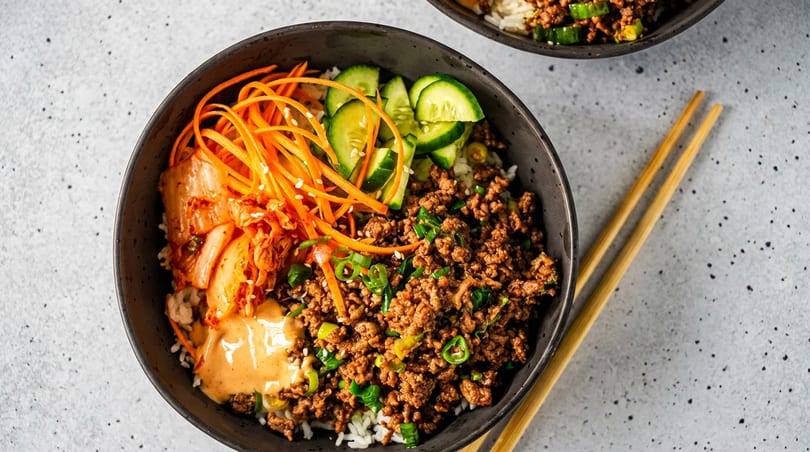 A Korean beef bulgogi bowl with rice, kimchi, carrots, cucumbers, and spicy sauce, with chopsticks.