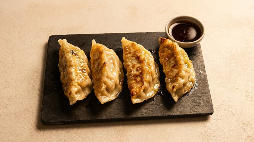 Four golden-brown pan-fried dumplings with a small bowl of dark dipping sauce on a black slate.