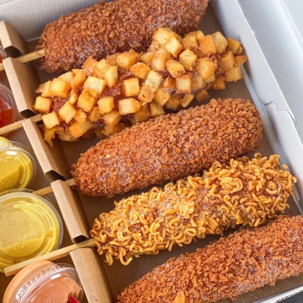 Assortment of Korean corn dogs with panko, diced potato, and ramen noodle toppings in a takeout box with sauces.