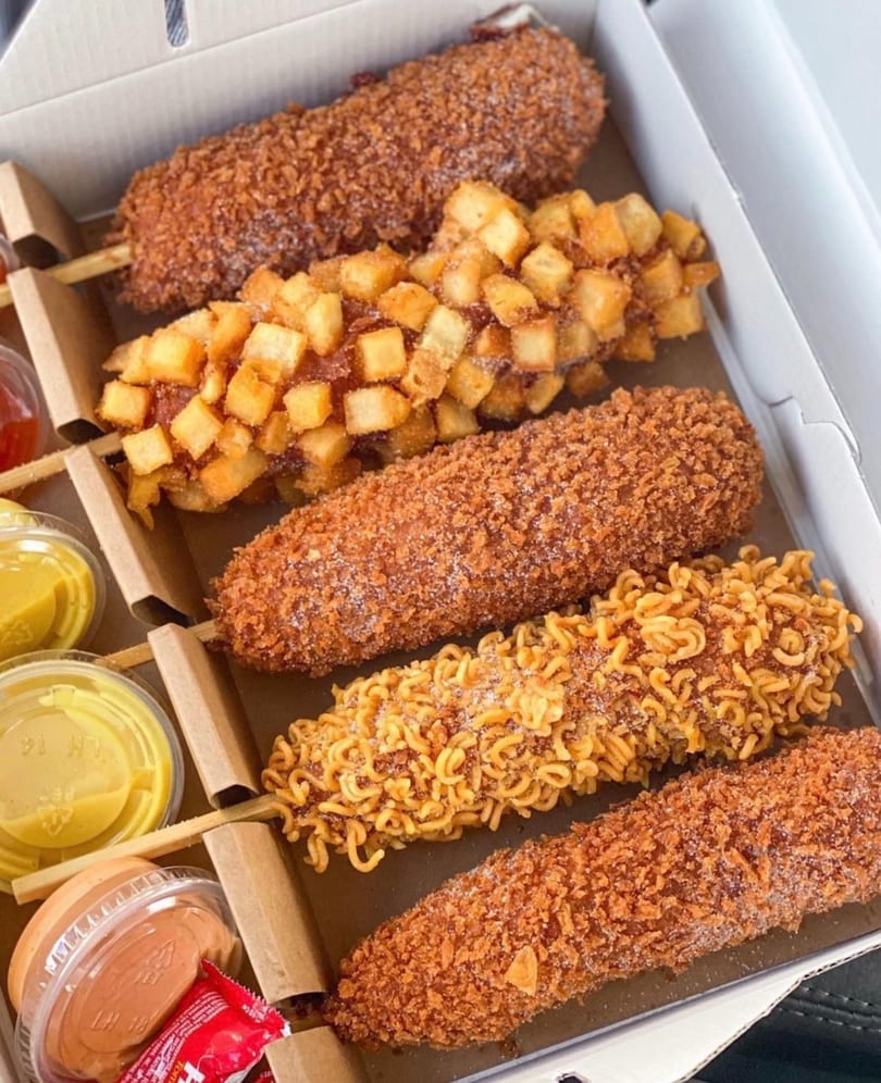 Assortment of Korean corn dogs with panko, diced potato, and ramen noodle toppings in a takeout box with sauces.