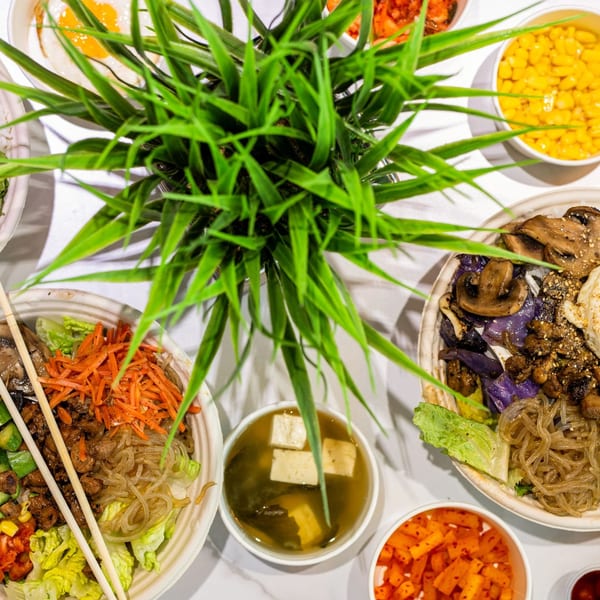 Multiple vibrant Asian food bowls with rice, vegetables, and proteins, arranged around a green plant.