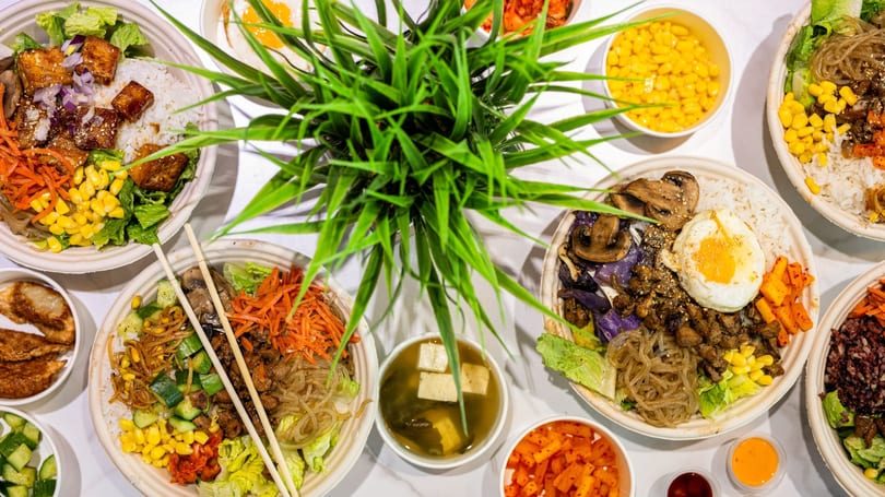 Multiple vibrant Asian food bowls with rice, vegetables, and proteins, arranged around a green plant.