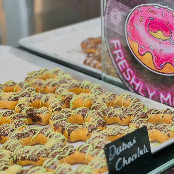 Tray of "Dubai Chocolate" donuts with chocolate, green icing, and shredded topping, near a donut sign.