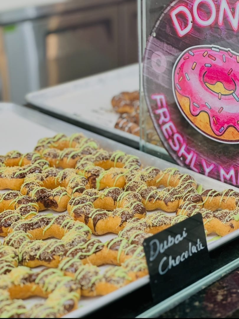 Tray of "Dubai Chocolate" donuts with chocolate, green icing, and shredded topping, near a donut sign.